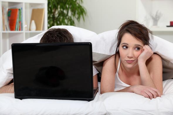 Junger Mann vor seinem Laptop im Bett, die Freundin liegt gelangweilt daneben.