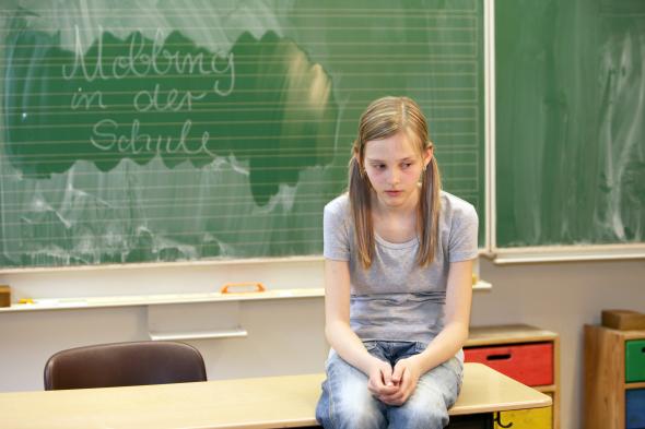Schülerin sitzt auf einem Tisch - im Hintergrund steht an der Tafel "Mobbing in der Schule"