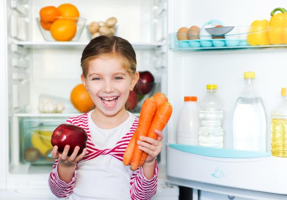 Kleines Mädchen steht vor einem Kühlschrank, und hält in der einen Hand einen Apfel und in der anderen Karotten.
