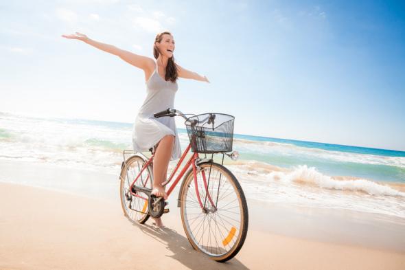 Junge Frau fährt freihändisch und glücklich auf dem Fahrrad am Strand entlang.